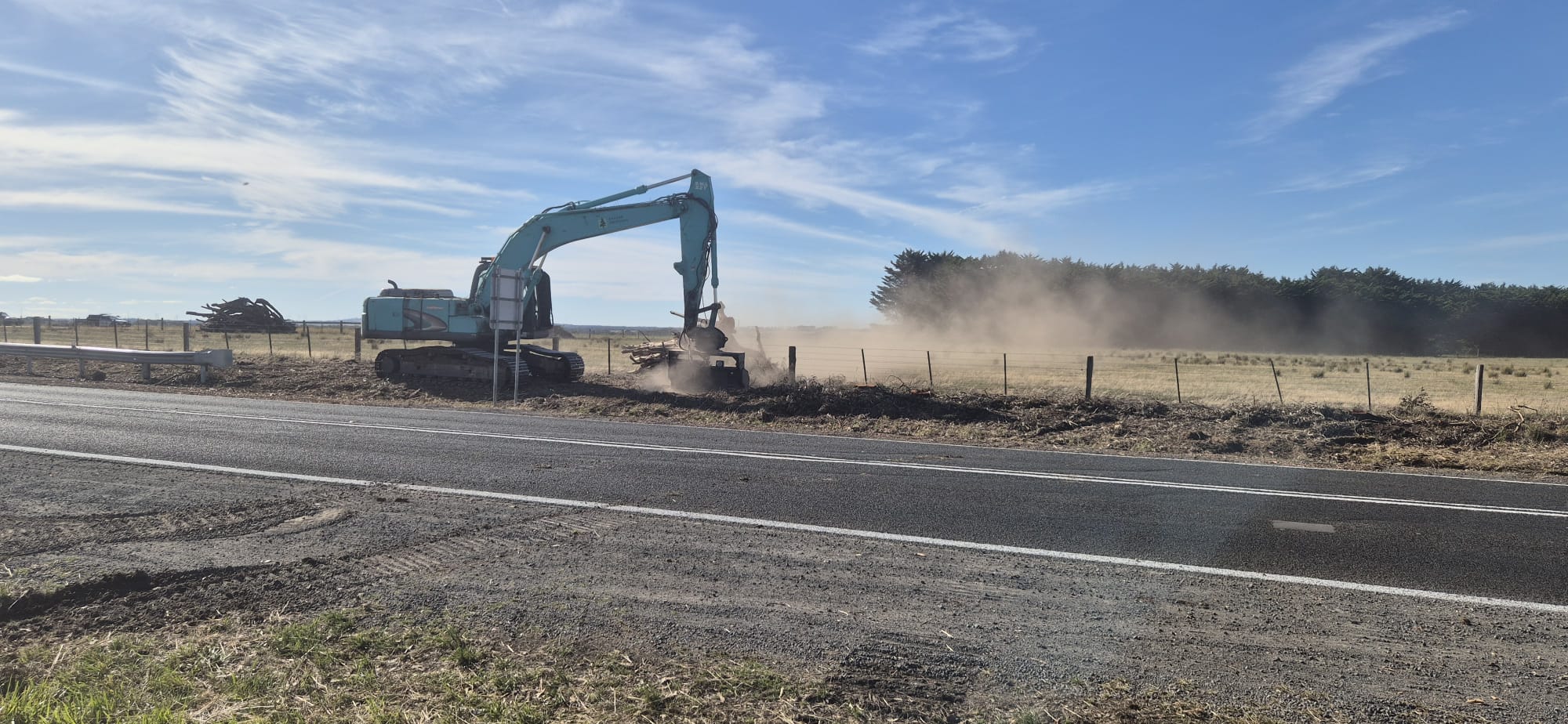 Excavator with mulching head clearing roadside trees beside live traffic