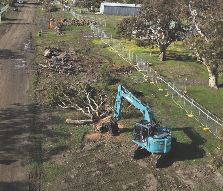 Aerial view of a blue excavator clearing trees along a fenced roadside site